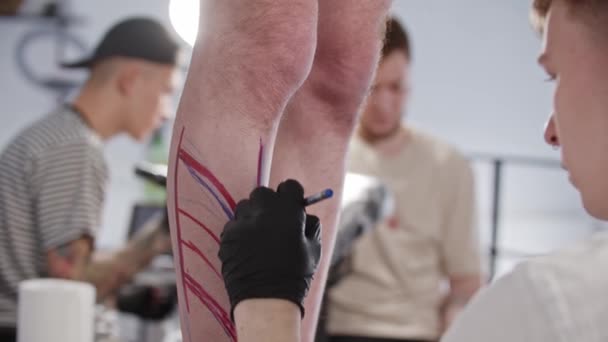 Jeune homme tatoueur dessiner un croquis sur la jambe sur son client avec un stylo bleu — Video