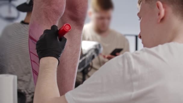Mladý muž tetování umělec kreslit náčrt na nohu na jeho klienta s červenou značkou v salonu — Stock video
