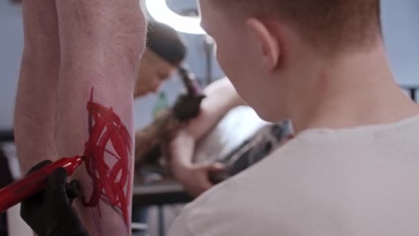 Jongeman tatoeage kunstenaar tekening een schets op het been op zijn cliënt met een rode markering door zijn hand — Stockvideo