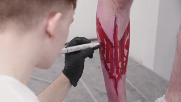 Jongeman tatoeage kunstenaar tekening een schets met letters op het been op zijn cliënt met een rode marker — Stockvideo