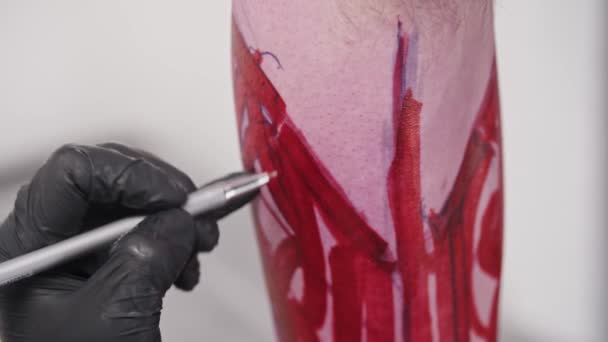若い男の入れ墨のアーティストは、赤いマーカーの上にペンで彼のクライアント上の足の上に手紙でスケッチを描く — ストック動画
