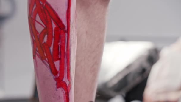 纹身艺术家在草图上添加了最后的润色，在他的客户的腿上有字母，并在红记号上涂上了笔 — 图库视频影像