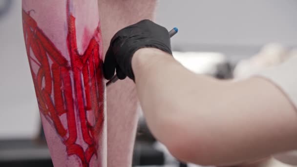 Tattoo kunstenaar toe te voegen afwerking raakt aan een schets op het been van zijn cliënt met een pen over de rode marker — Stockvideo