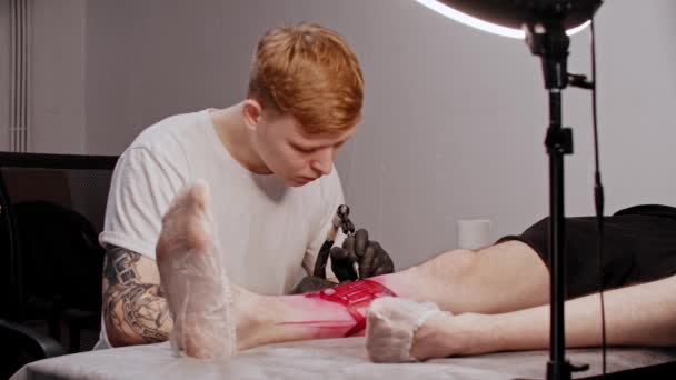 Tattoo salon - young man tattooing the contour on the leg — Stock Video