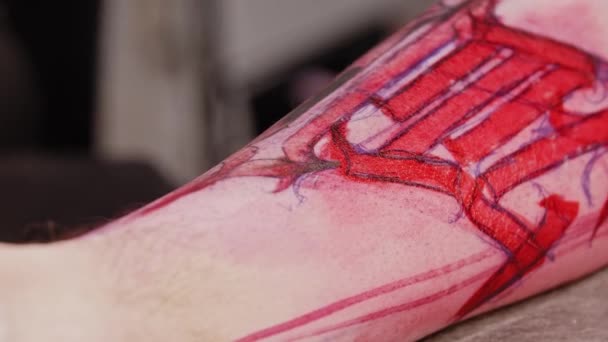 Tatueringssalong - ung man gör en tatuering på benet av sin klient över en röd skiss — Stockvideo