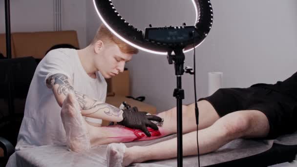 Dövme salonu - genç adam halka ışığının altındaki bacağa dövme yapıyor. — Stok video
