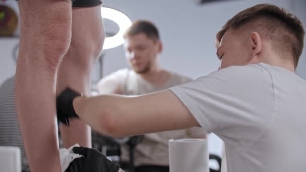 Mladý muž holí nohu svého klienta pro tetování v tetovacím salonu — Stock video