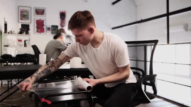 Hombre tatuado maestro del tatuaje preparando el lugar para su trabajo - cubriendo la mesa con polietileno — Vídeo de stock