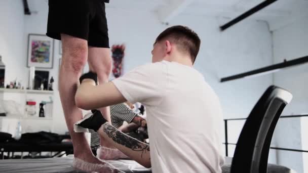 Salón de tatuajes - joven afeitándose la parte inferior de la pierna de su cliente — Vídeos de Stock