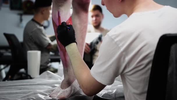 纹身美容院（Tattoo salon）-年轻的纹身艺术家，手拿着红色记号在他的客户的腿上画了一幅草图 — 图库视频影像