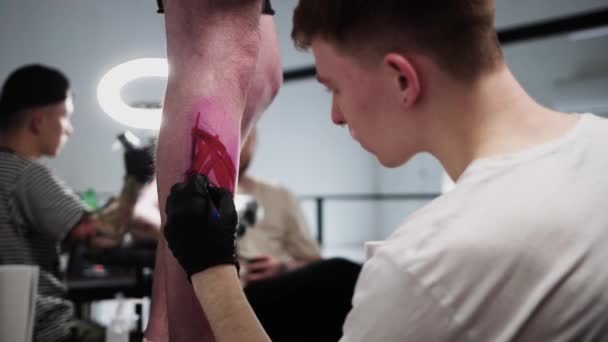 Tattoo salon - jonge man tatoeage kunstenaar tekening van een schets op het been op zijn cliënt met een rode marker en een pen door zijn hand — Stockvideo