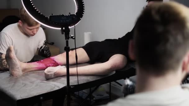 Salão de tatuagem - jovem mestre tatuando letras grandes na perna — Vídeo de Stock