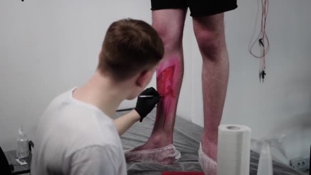 Tattoo studio -若い男のマスターは、赤い明るいマーカーで彼のクライアント上の足にスケッチ文字を描く — ストック動画