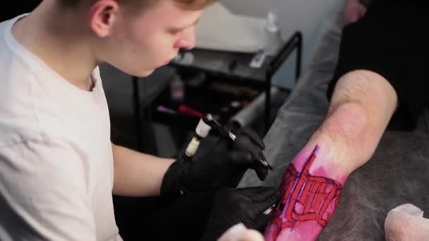 Salão de tatuagem - jovem mestre tatuagem letras na perna sobre marcador vermelho — Vídeo de Stock