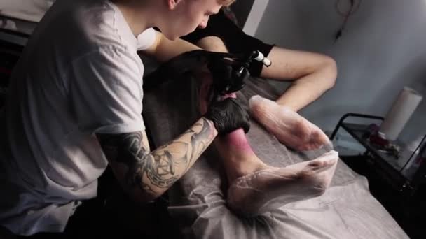 Salon de tatouage - client masculin allongé sur le canapé et se faisant tatouer la jambe en style graffiti - le maître remplit le tatouage d'encre noire — Video