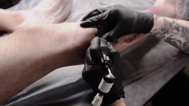 Salão de tatuagem - o cliente deitado no sofá e recebendo uma tatuagem na perna em estilo grafite - o mestre enchendo a tatuagem com tinta preta — Vídeo de Stock