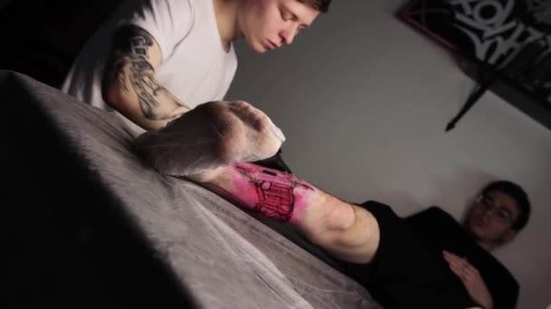 Tattoo salon - male client lying on the couch and getting a tattoo on his leg - the master filling the tattoo with black ink — Stock Video