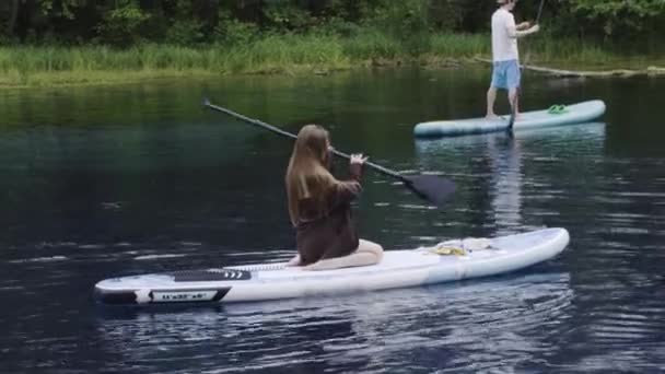 Ung brunett kvinna sitter på knä på brädorna i floden och vrider paddeln över huvudet — Stockvideo