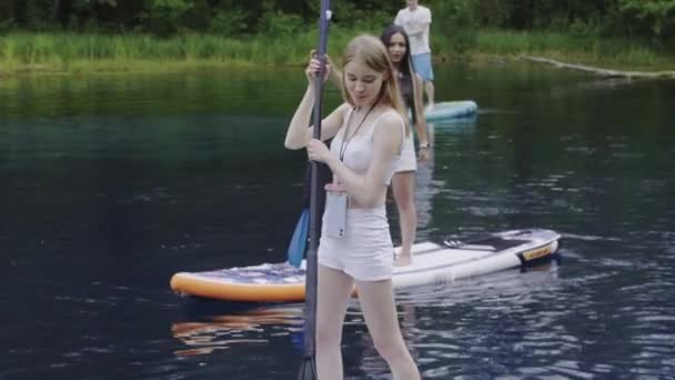Młoda blondynka piękna kobieta stojąca na desce nad rzeką i używająca wiosła — Wideo stockowe