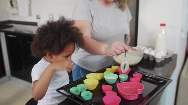 Zwart klein meisje kijken naar haar wit moeder gieten deeg in kleine vormen — Stockvideo