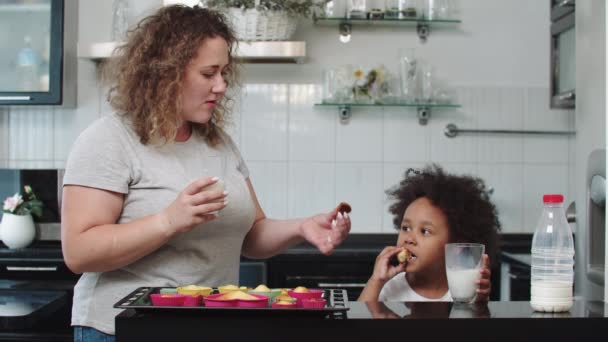 Vit mamma med sin svarta blandade dotter äter muffins med mjölk i köket — Stockvideo