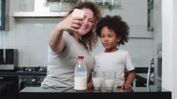 Madre blanca y su hija negra mezclada tomando una selfie en el teléfono - vasos de leche sobre la mesa — Vídeos de Stock