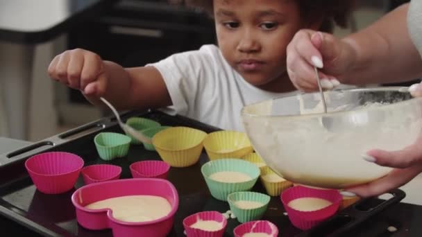 Uma menina negra e sua mãe branca derramando massa líquida crua nos moldes de bolo com uma colher — Vídeo de Stock