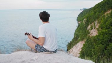 Genç sakin adam uçurumda otururken ukulele çalıyor.