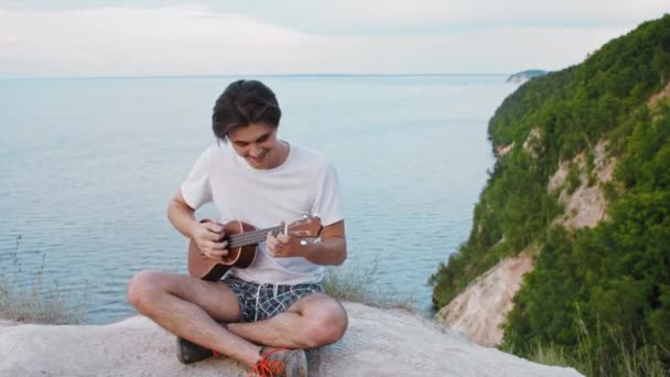 Jeune homme souriant jouant ukulélé assis sur la montagne — Video