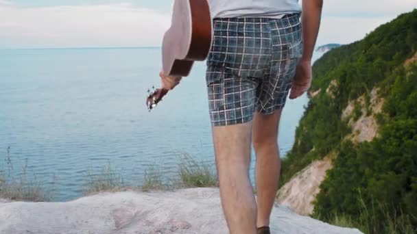 Молодой человек идет к утесу и начинает играть на укулеле — стоковое видео