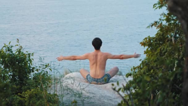 Egy fiatal félmeztelen férfi meditál egy sziklán kora reggel. — Stock videók