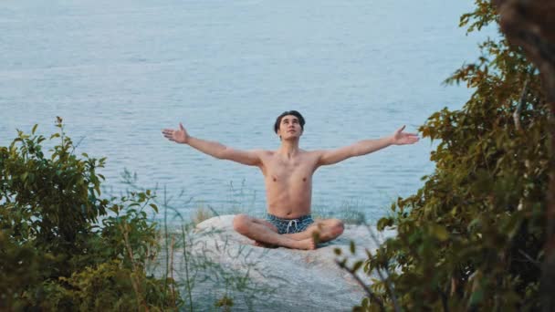 Un joven sin camisa medita en una montaña por la mañana temprano mirando a la cámara — Vídeos de Stock
