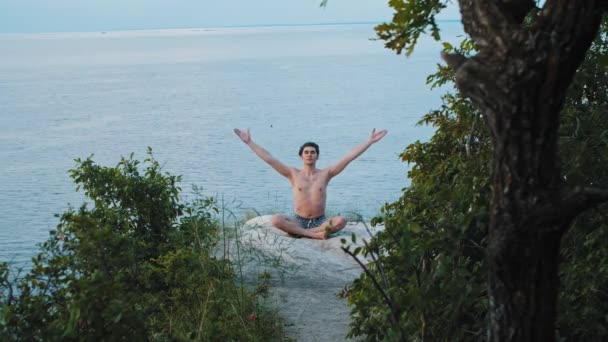 Un joven sin camisa medita en una montaña sentado hacia la cámara — Vídeos de Stock