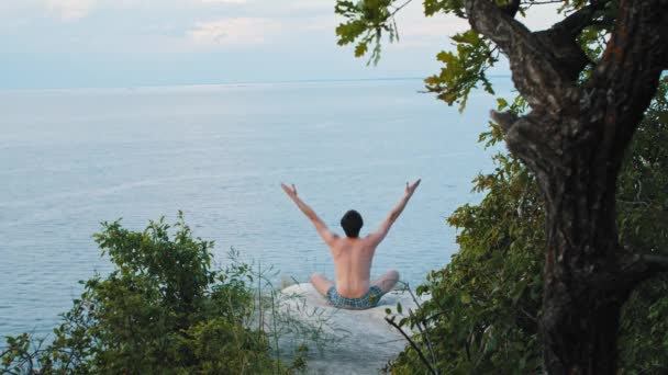 Een jonge shirtloze man mediteert op een klif in de vroege bewolkte ochtend — Stockvideo