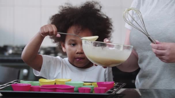 Une petite fille noire drôle versant de la pâte liquide crue dans les moules à gâteau avec une cuillère — Video