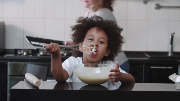 Cuisson familiale mixte - petite fille noire mangeant de la pâte liquide crue du fouet et sa mère apporte les moules à gâteau — Video