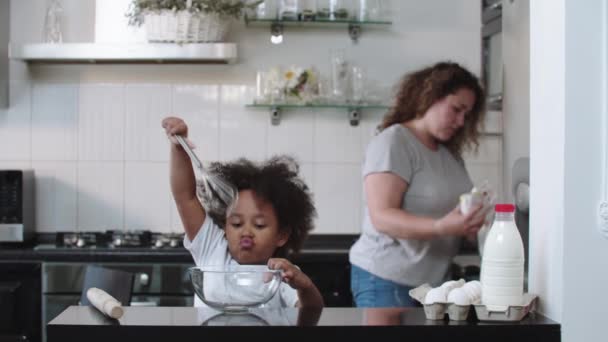 Little black girl playing with a whisk in a glass bowl while her mother washing dishes — Stock Video