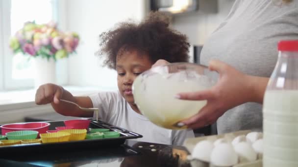 Familjebakning - svart liten flicka och hennes mamma häller flytande deg i små kakformar — Stockvideo