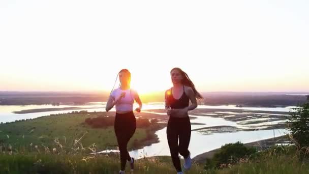 Dos mujeres jóvenes en ropa deportiva corriendo en el campo al atardecer brillante — Vídeos de Stock