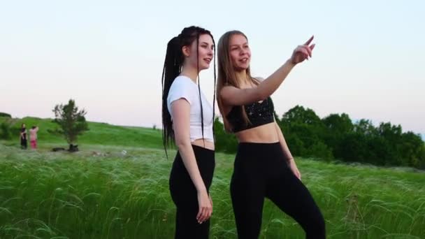 Dos mujeres jóvenes de pie en la hierba alta y mirando el atardecer apuntando al cielo — Vídeos de Stock
