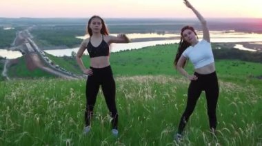 İki genç kadın yeşil bir tarlada durup egzersizleri ısıtıyorlar.