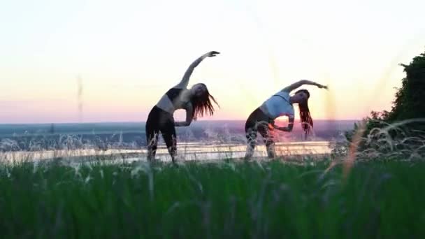 Dos mujeres jóvenes de pie en un campo verde y haciendo ejercicios de calentamiento - doblando a los lados en eraly puesta de sol — Vídeos de Stock