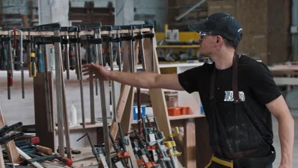 El hombre en un taller de carpintería toma un instrumento mirando a la cámara — Vídeos de Stock