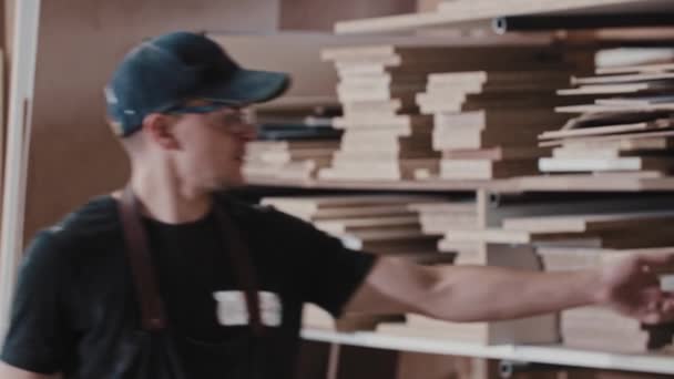 Homem em uma oficina de carpintaria leva uma peça de madeira da prateleira e soprando poeira a partir dele olhando para a câmera — Vídeo de Stock