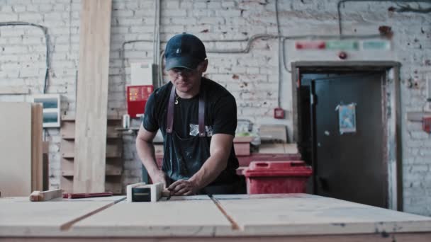 Homem em uma oficina de carpintaria cortando pedaços de um pequeno detalhe de madeira usando uma máquina de corte estática — Vídeo de Stock