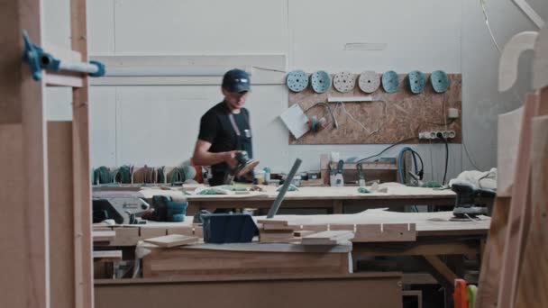 Man in een timmermanswerkplaats brengt een houten stuk binnen en staat op het punt het te malen. — Stockvideo