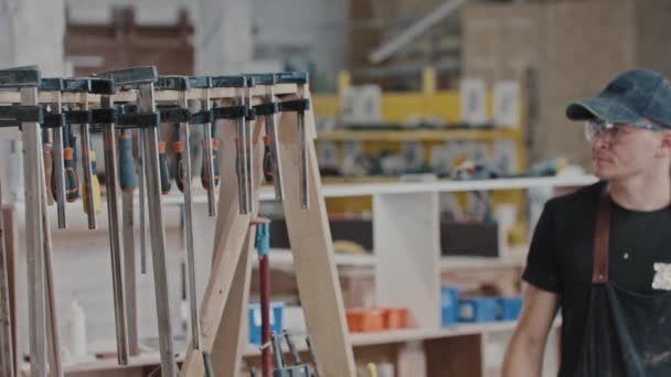 El hombre camina en un taller de carpintería y toma un instrumento mirando a la cámara — Vídeos de Stock