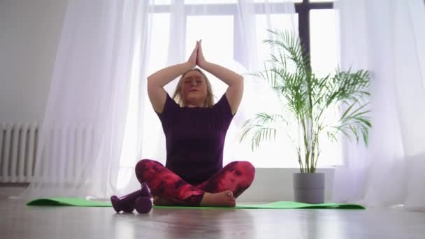 Yoga en interiores - mujer rubia con sobrepeso sonriente meditando en estera de yoga en estudio moderno blanco — Vídeo de stock