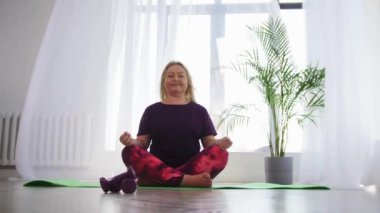Yoga kapalı alanda - beyaz modern odada yoga minderi üzerinde meditasyon yapan şişman sarışın kadın.