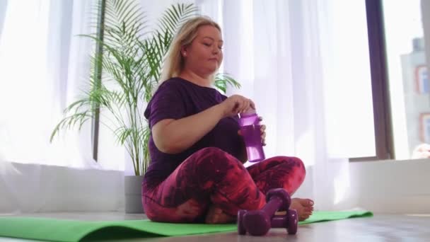 Фитнес-тренировка - блондинка с избыточным весом сидит на коврике для йоги и пьет воду из бутылки — стоковое видео